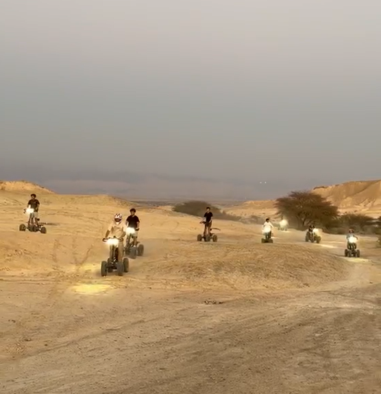 שיירת רכבי איזי ריידר עם אורות דולקים נוסעת בשביל חול בלב מדבר הערבה בשעת שקיעה
